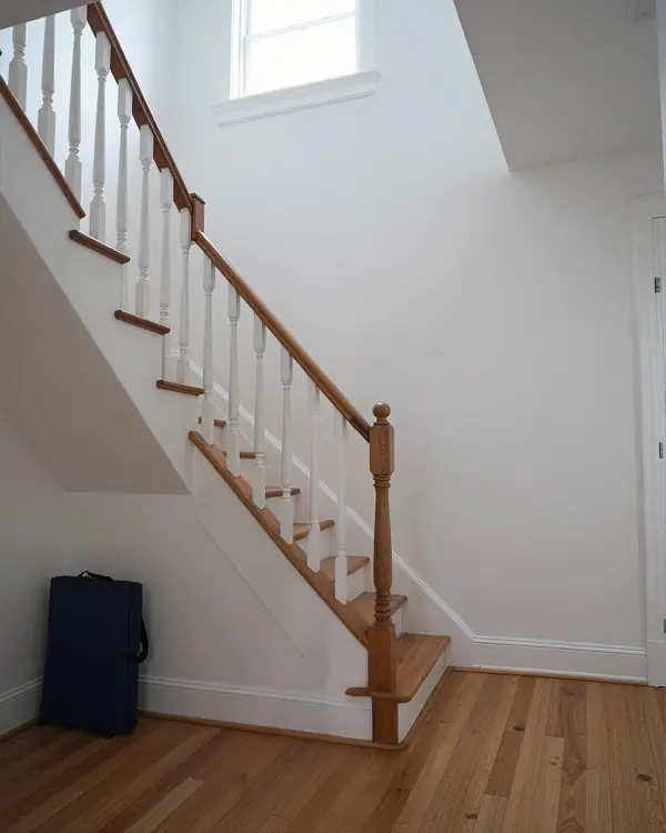 The foot of a staircase in a house, white walls and a wood floor.